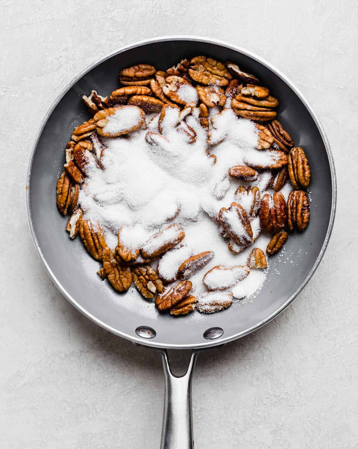 Pecan halves in a gray skillet with granulated sugar over the pecans.