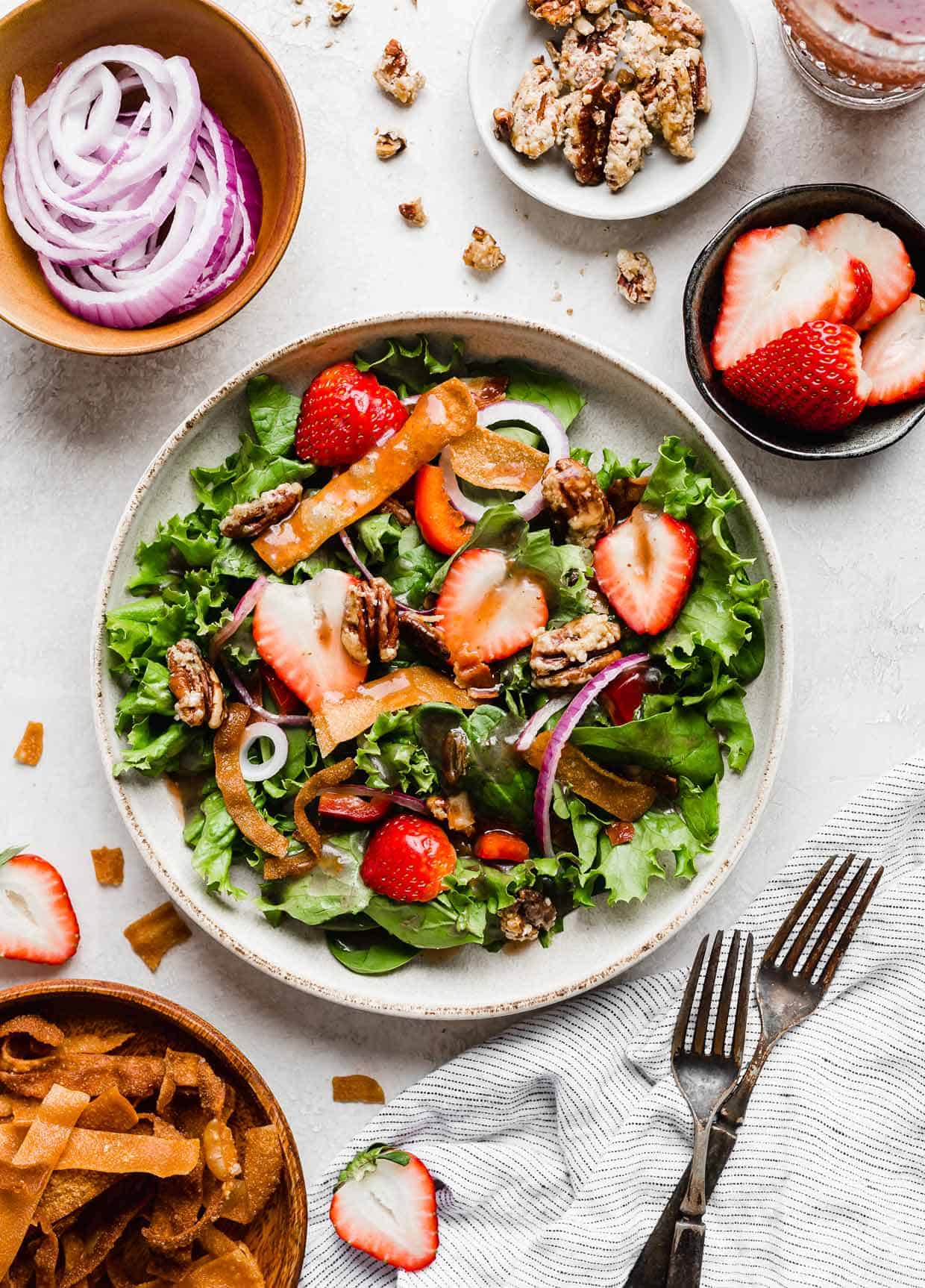Overhead photo of a strawberry spinach salad on a white background with two forks, strawberries, fried wontons, and red onions scattered around the salad.
