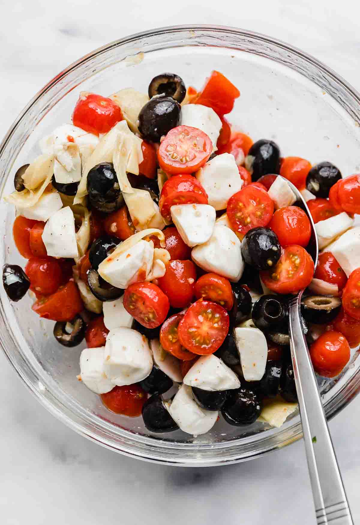 A glass bowl filled with Mozzarella, Olive, and Tomato Artichoke Salad in it on a white background.