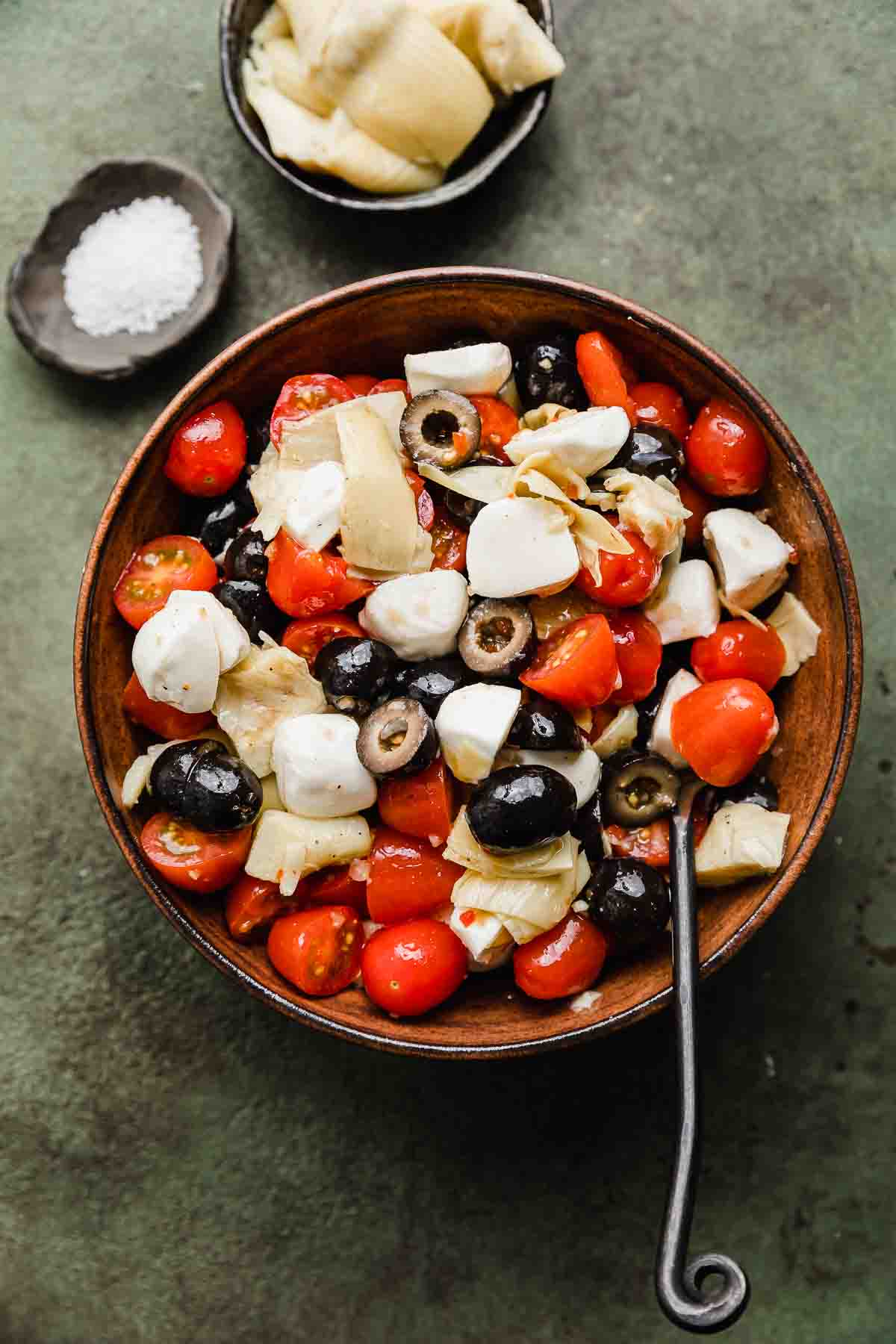 A brown bowl filled with Tomato Artichoke Salad with olives and mozzarella cheese balls, on a forest green colored background.