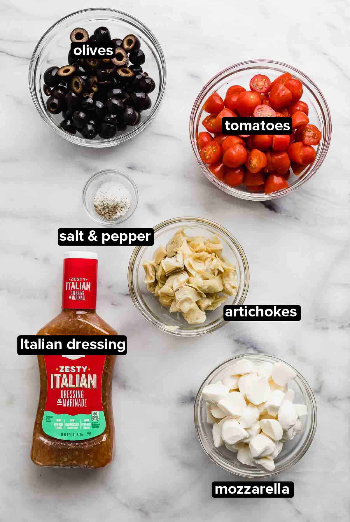 Tomato Artichoke Salad ingredients on a white background, ingredients include olives, Italian dressing, mozzarella pearls, tomatoes, artichokes, salt and pepper.