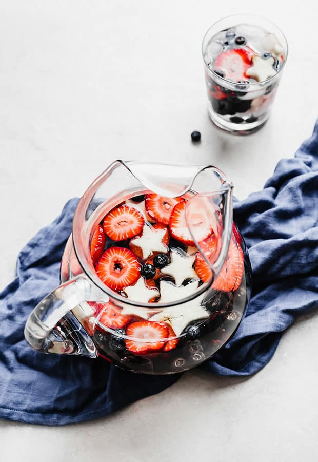 A glass pitcher full of sliced strawberries, blueberries, blackberries, and star shaped apples.