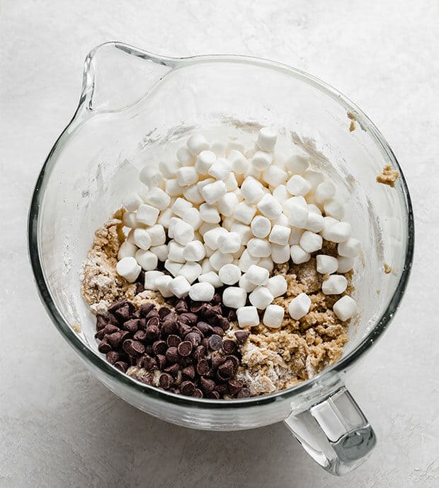 A glass mixing bowl with cookie dough batter, marshmallows, and chocolate chips.
