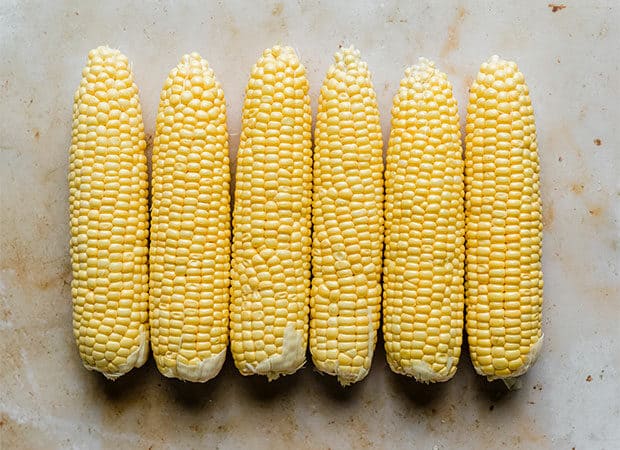 Six husked corn on the cob against a tan background.