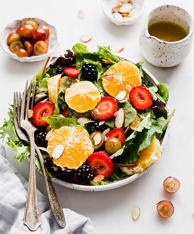A large plate full of mixed salad greens, topped with sliced oranges, strawberries, grapes, and almonds.
