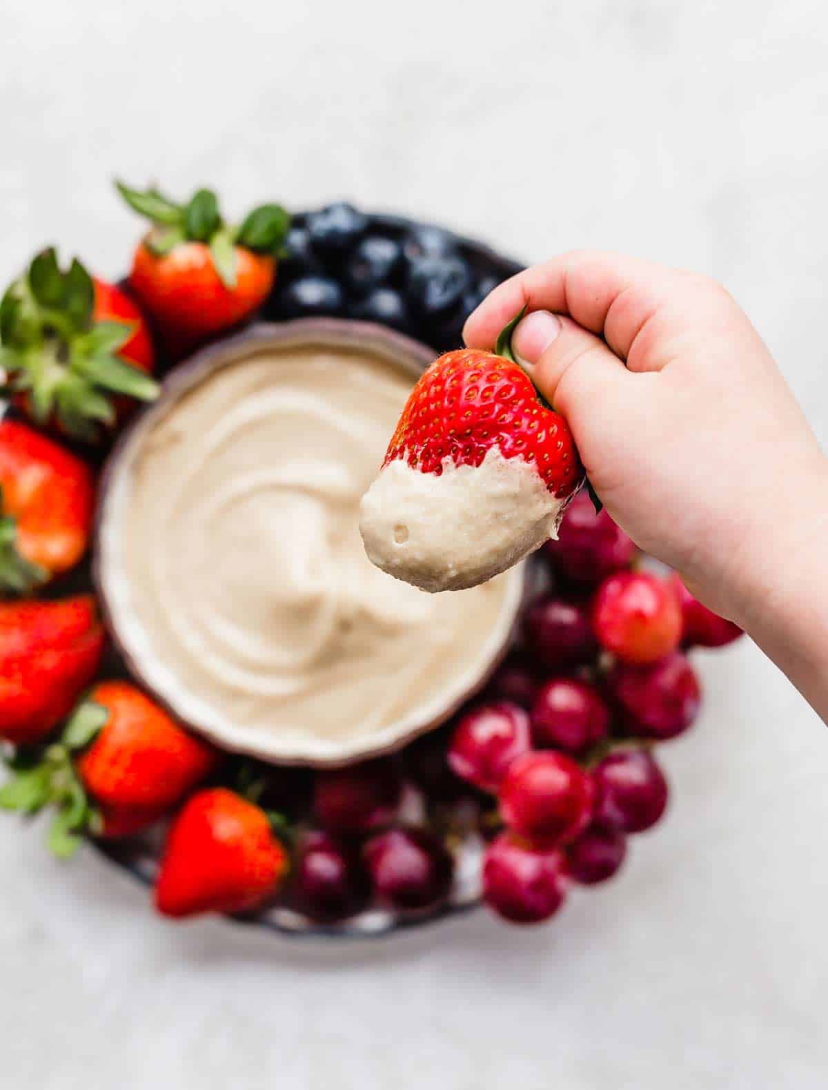 A hand dipping a strawberry into a tan colored Cream Cheese Fruit Dip.