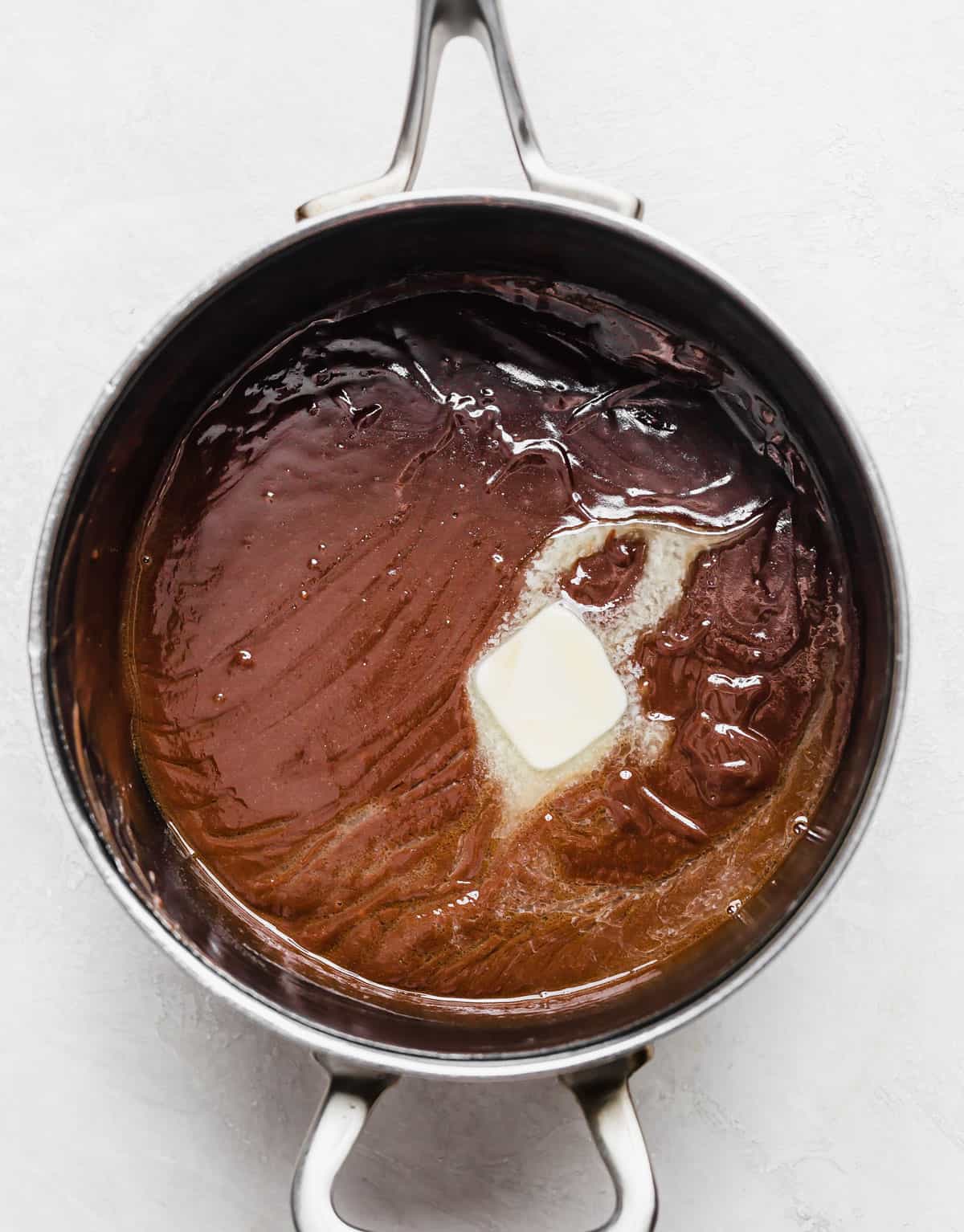 A tablespoon of butter melting into homemade hot fudge sauce in a saucepan.