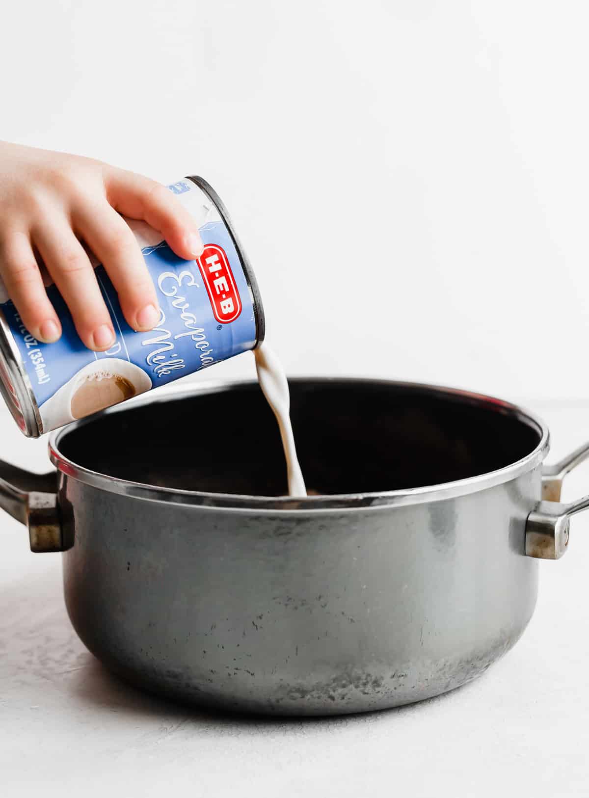 A hand pouring a can of evaporated milk into a gray saucepan.