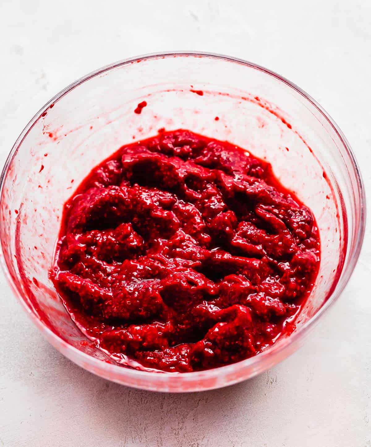 Mashed frozen raspberries to make freezer jam in a glass bowl on a white background.