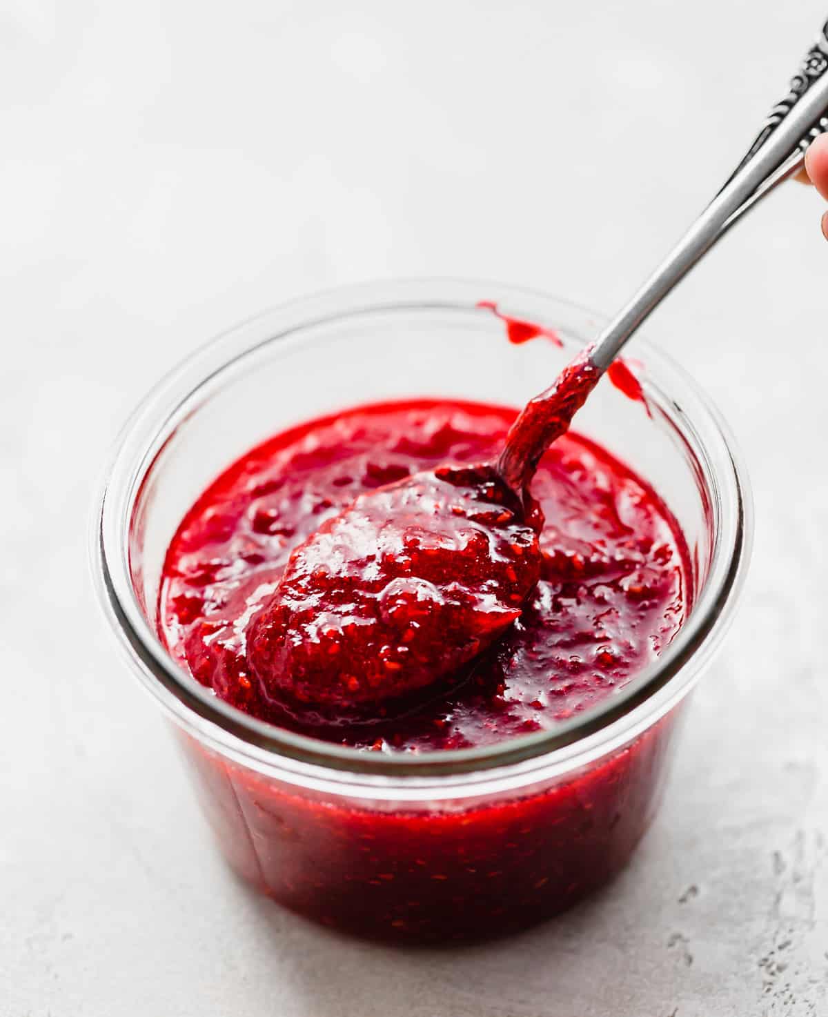 A glass jar filled with Raspberry Freezer Jam and a spoon scooping some out.