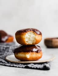 Two Boston Cream Donuts stacked on top of one another with the top donut having a bite taken out of it.