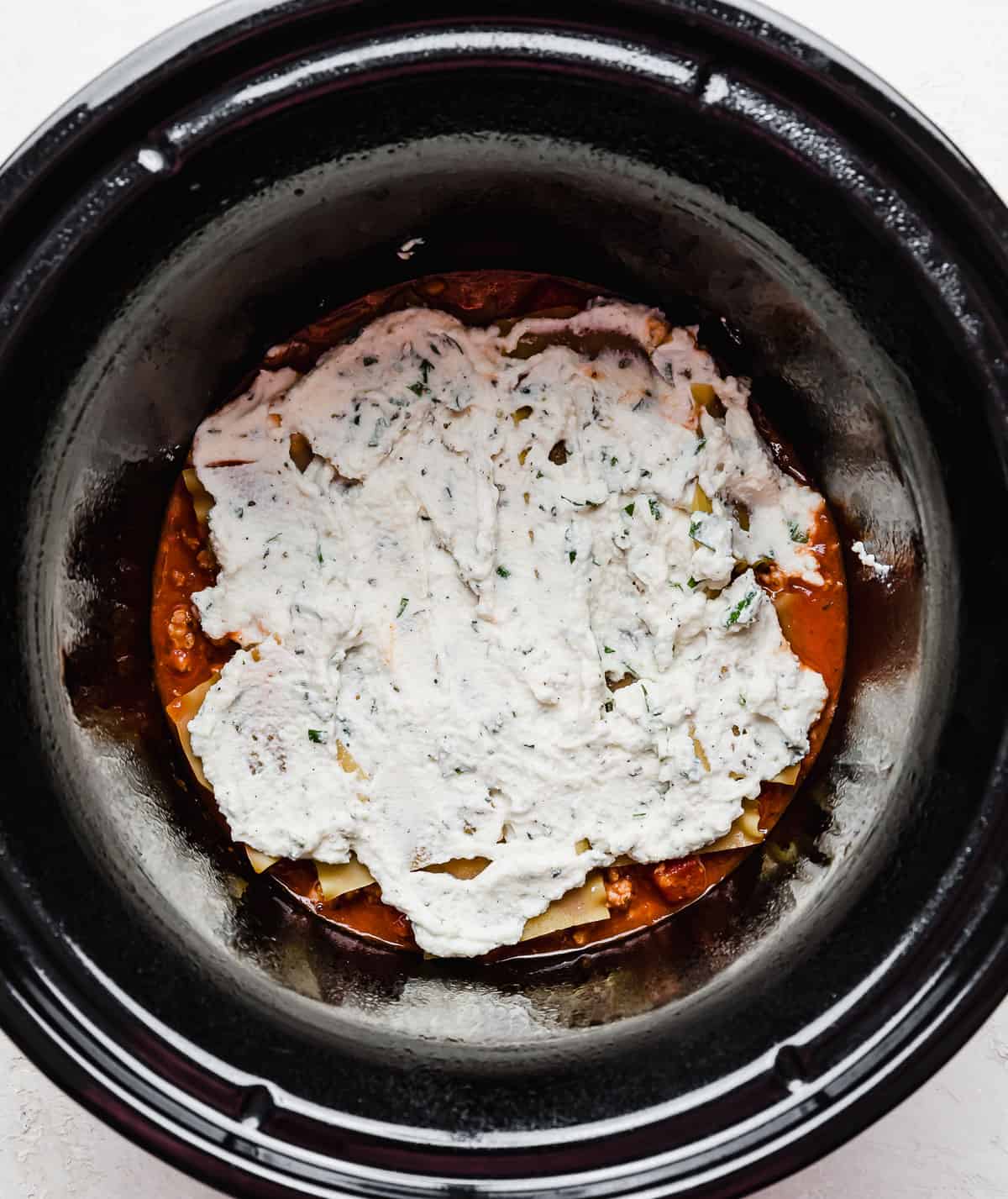 A round slow cooker with ricotta overtop lasagna noodles at the bottom of the crock pot.