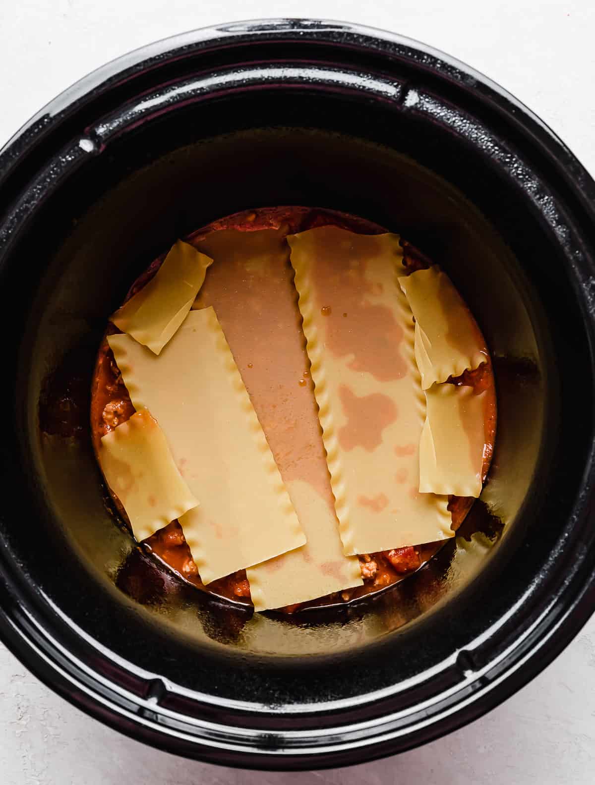 Lasagna noodles in the base of a black round slow cooker.