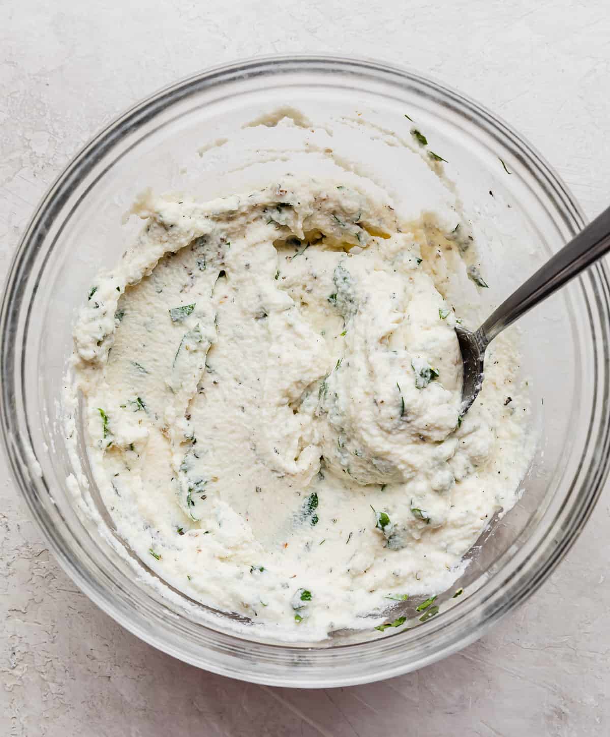A glass bowl with ricotta mixed with fresh parsley.