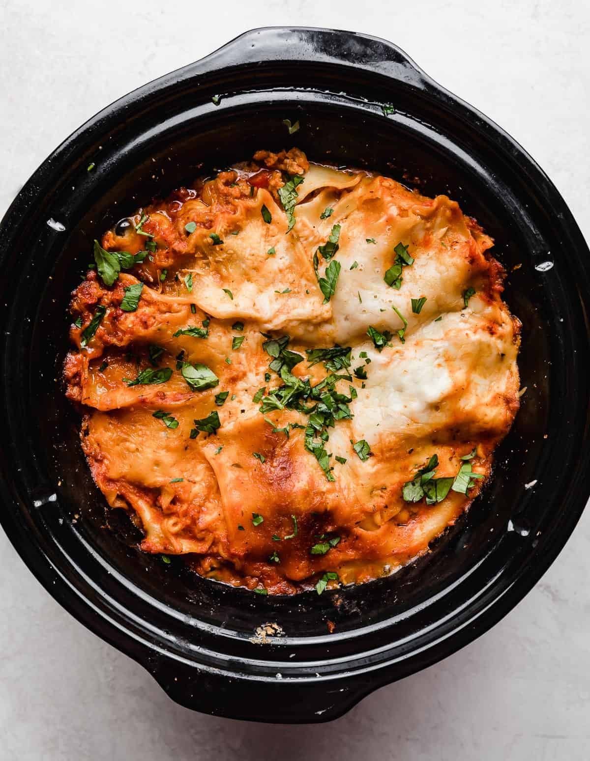 A warm freshly cooked Slow Cooker Lasagna in a black round crock pot garnished with fresh parsley.