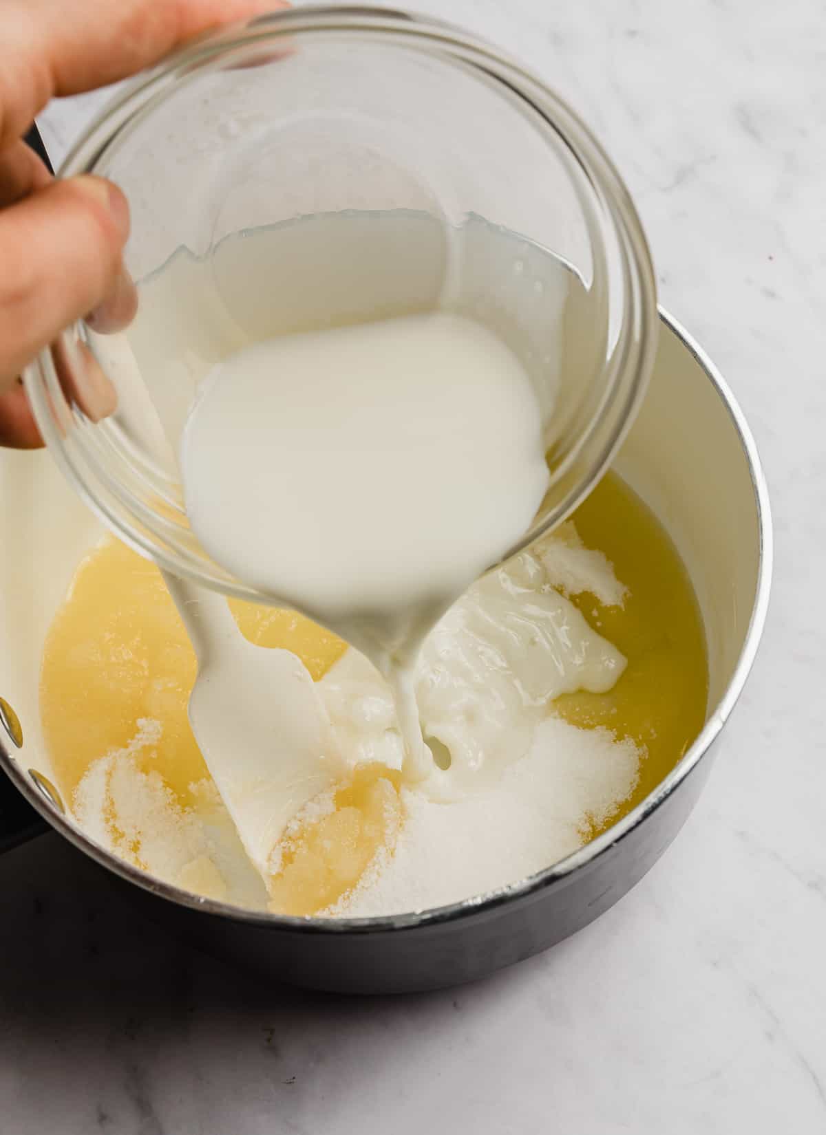 Milk being poured into a white pot that has melted butter and sugar in it.