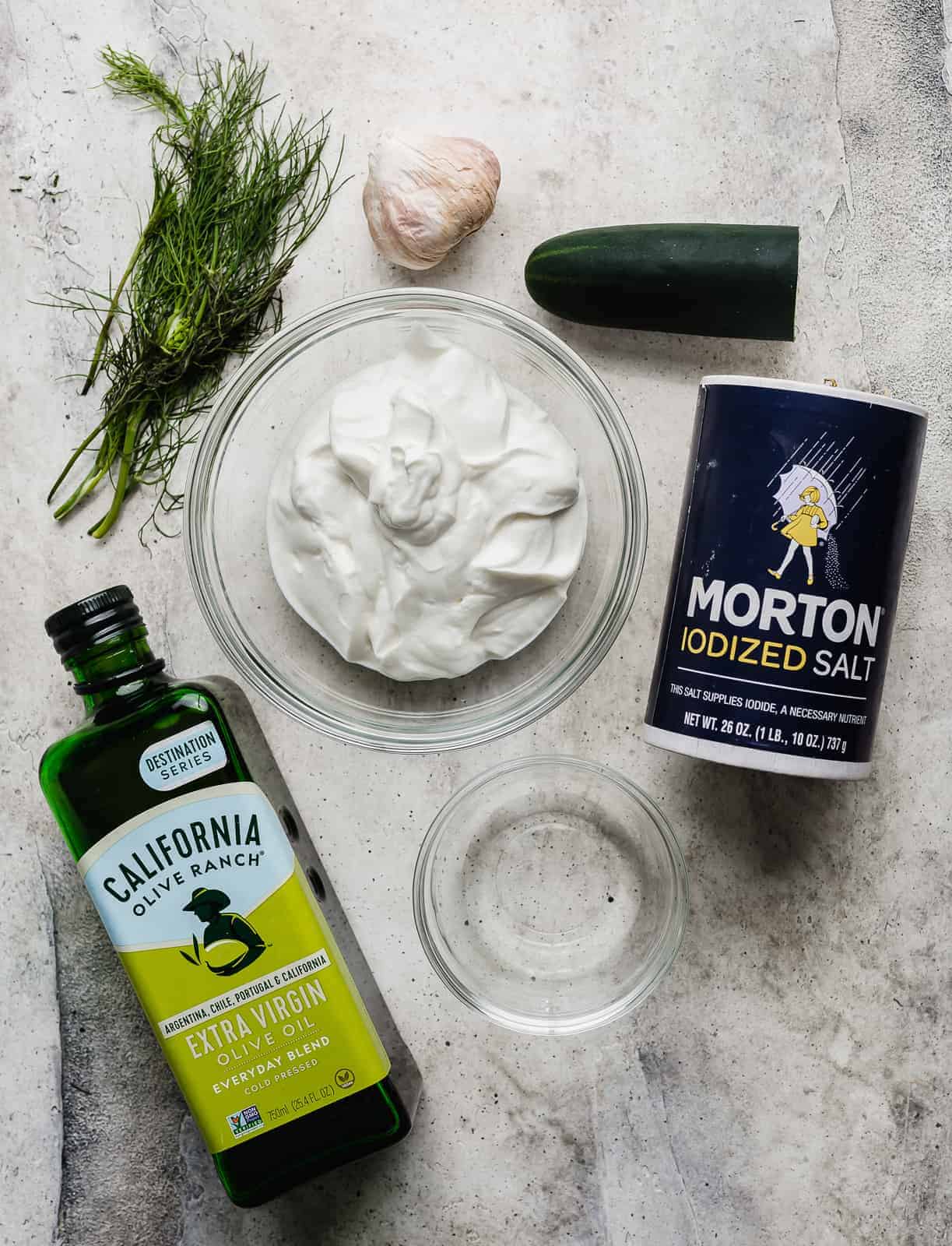 Tzatziki sauce ingredients: olive oil, dill, garlic, yogurt, salt, and cucumber on a grey background.