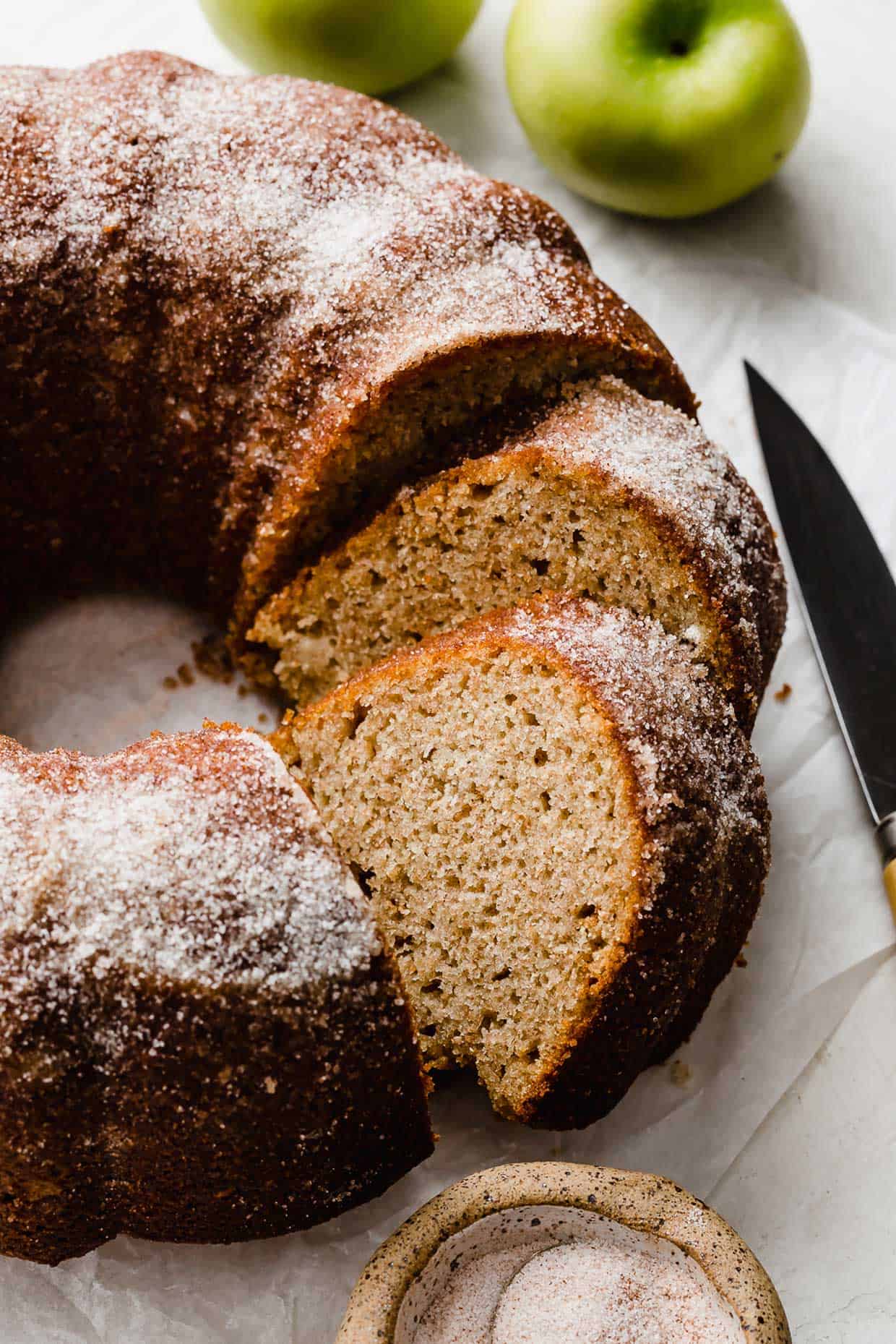An Apple Cider Bundt Cake topped with sugar with green apples in the background.
