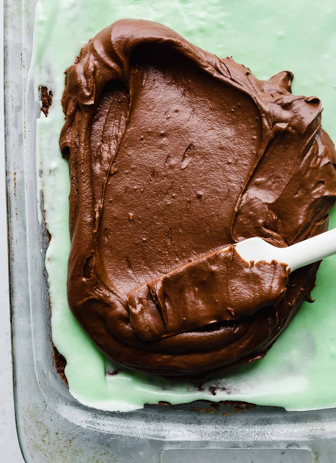 A spatula spreading chocolate frosting over mint frosting covered brownies.