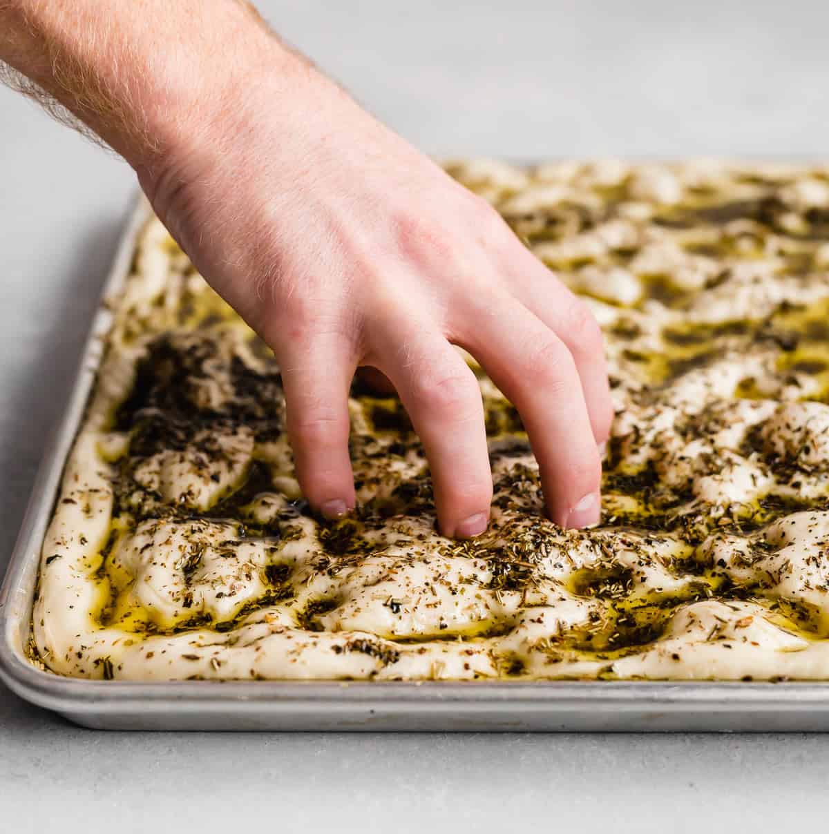 A hand pressing five fingers down into focaccia bread dough that's been topped with herb oil.