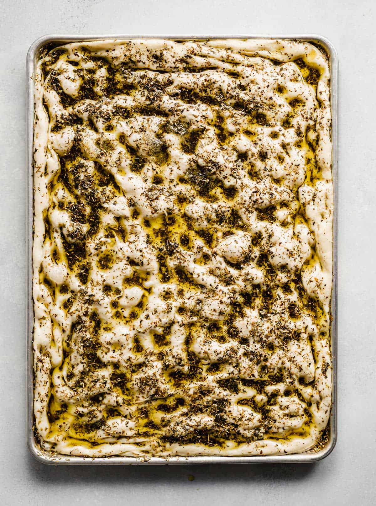 A baking sheet that has focaccia bread dough spread along the full size of the sheet, topped with herb oil.