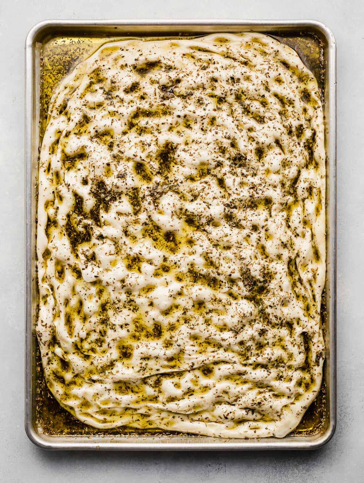 Raw focaccia bread dough dimpled and topped with herb oil on a baking sheet.