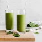 Two glasses full of a green protein smoothie on a wooden cutting board with a bowl of spinach in the background.