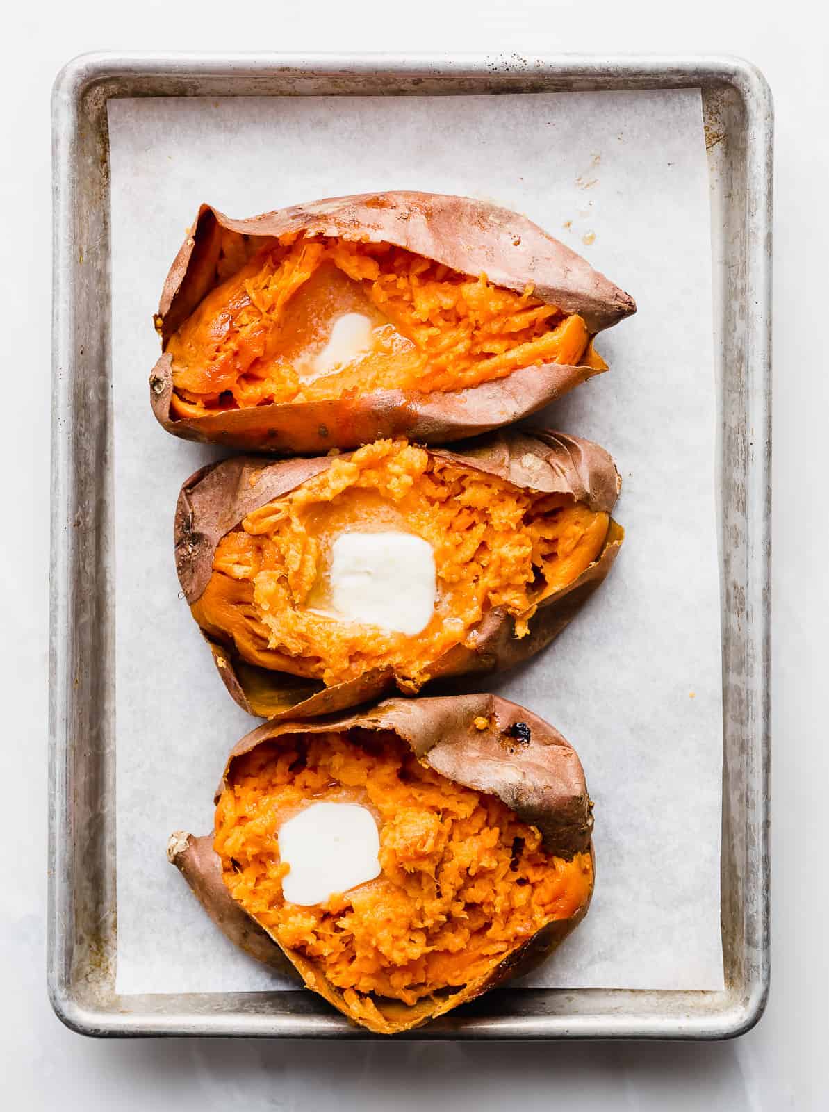 Three sweet potatoes on a parchment lined baking sheet, each cut open with a slice of butter melting in each potato.