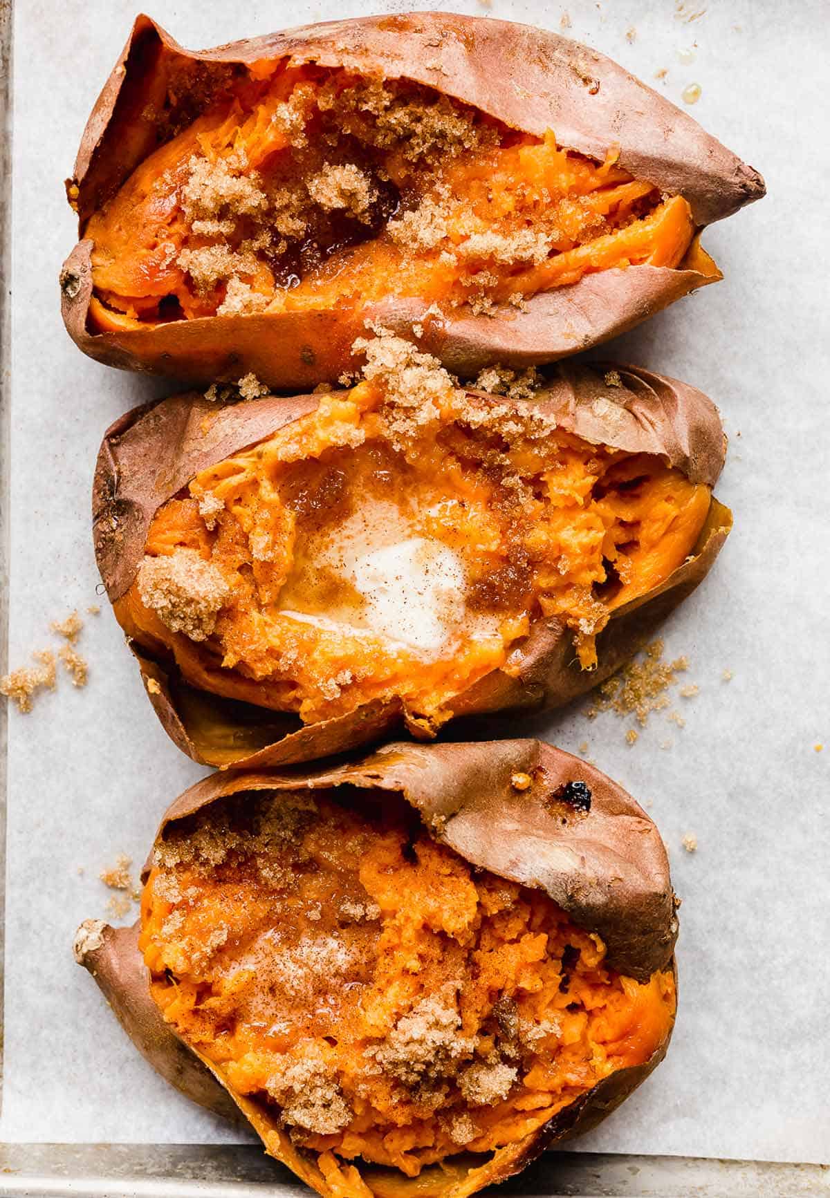 Three Baked Sweet Potatoes split open with melted butter and brown sugar sprinkled overtop.