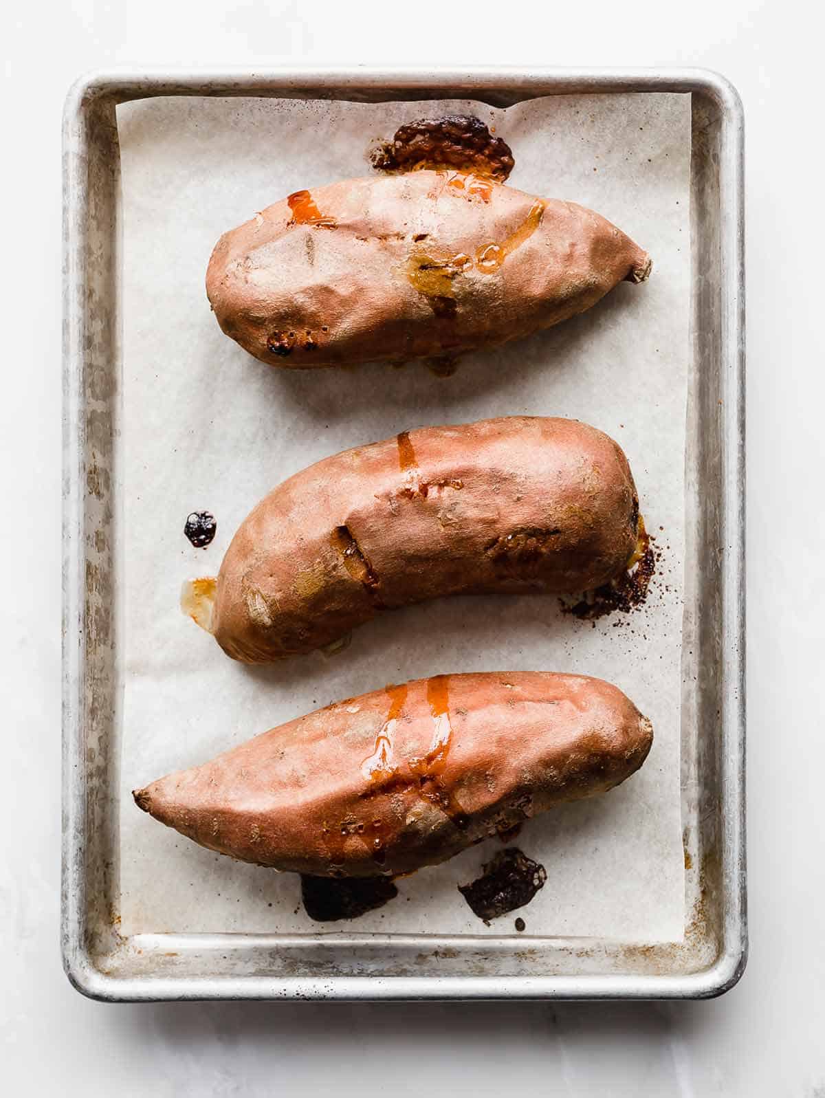 Three perfectly Baked Sweet Potatoes on a white parchment lined baking sheet.