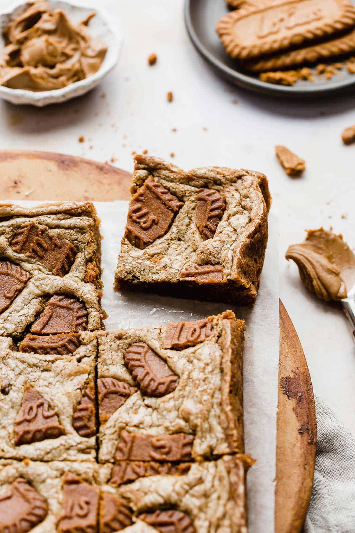 A square of Biscoff Brownies on a white parchment paper with Biscoff cookies in the background.