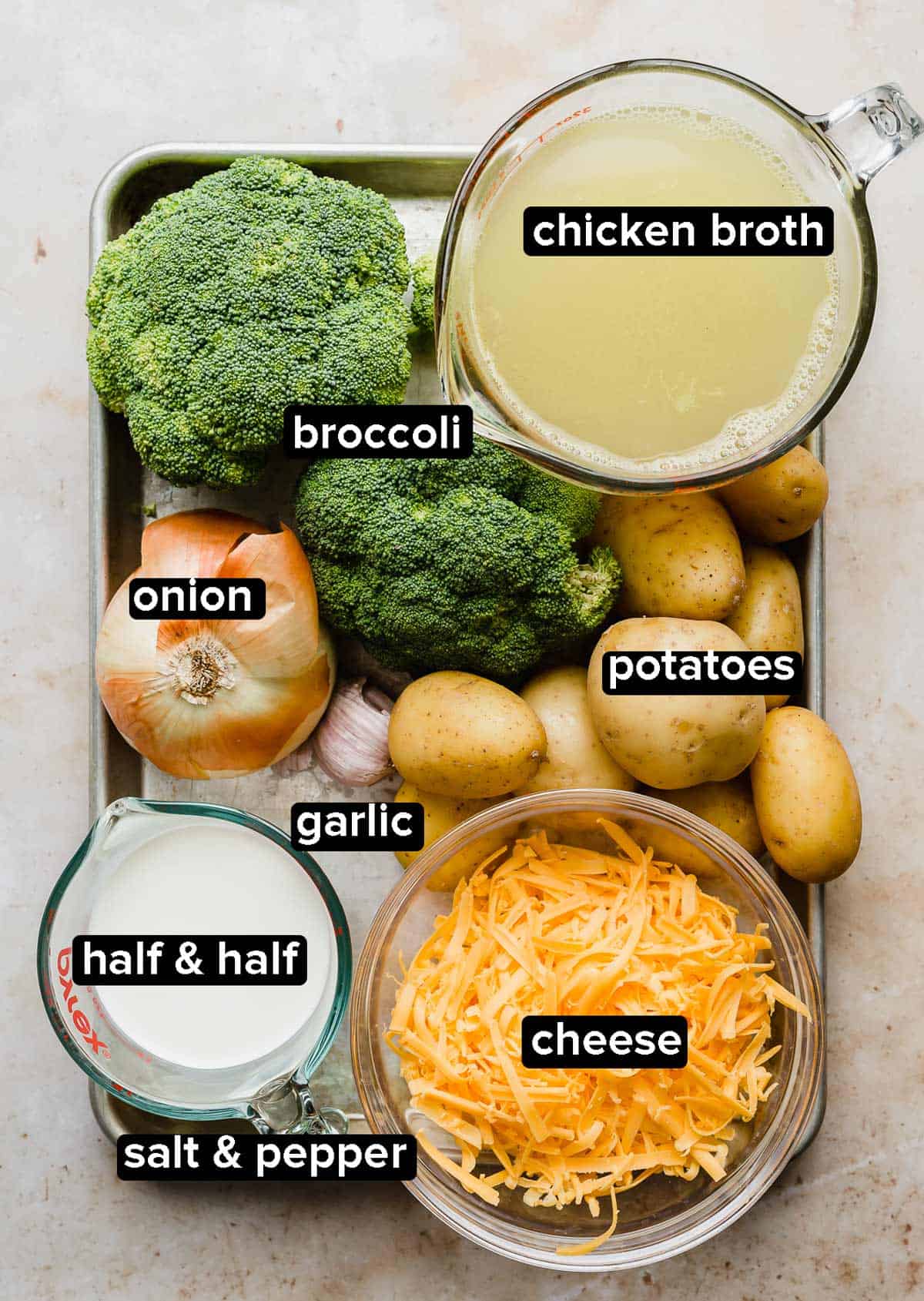 Broccoli Potato Soup ingredients on a metal baking sheet: broccoli, onion, potatoes, cheese, half and half, chicken broth, and garlic.