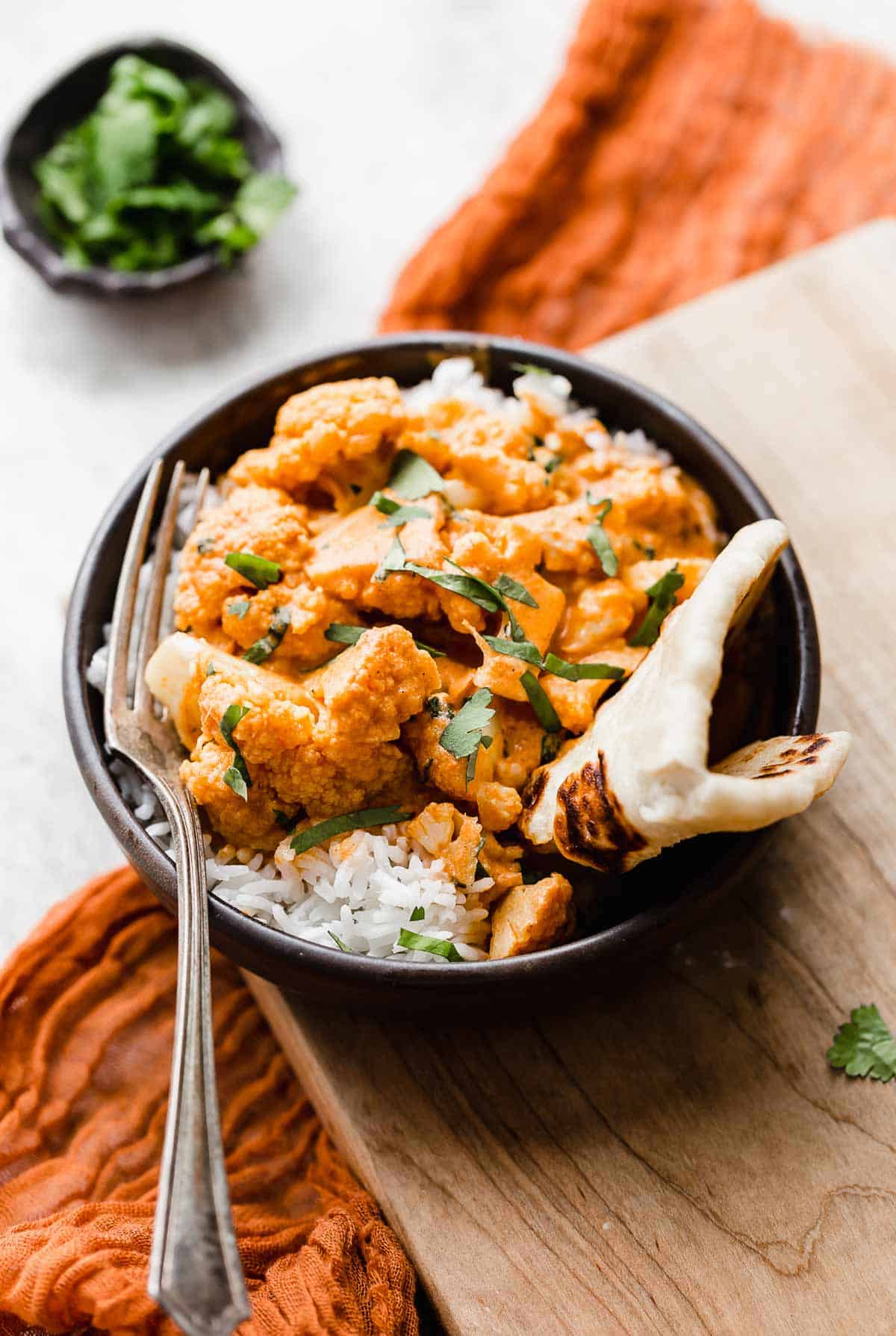 Red colored Indian Butter Cauliflower on a bed of white rice in a black bowl, with a piece of naan folded to the side and garnished with cilantro.