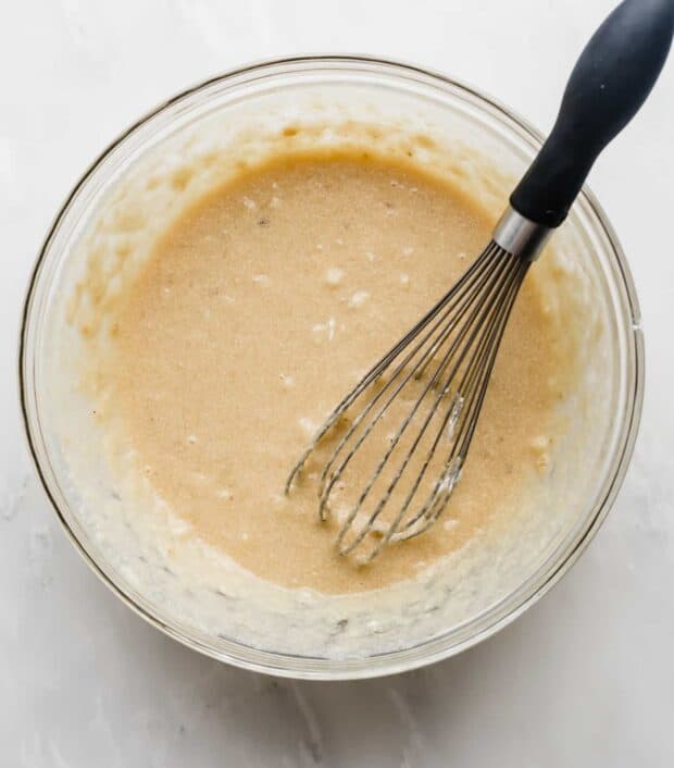 A whisk in a glass bowl stirring banana bread batter.
