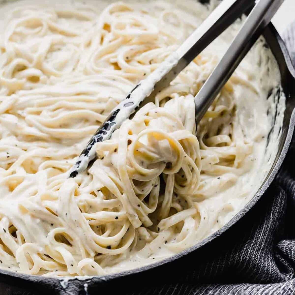 Creamy Fettuccine Alfredo in a black skillet with kitchen tongs being used to mix the creamy pasta.