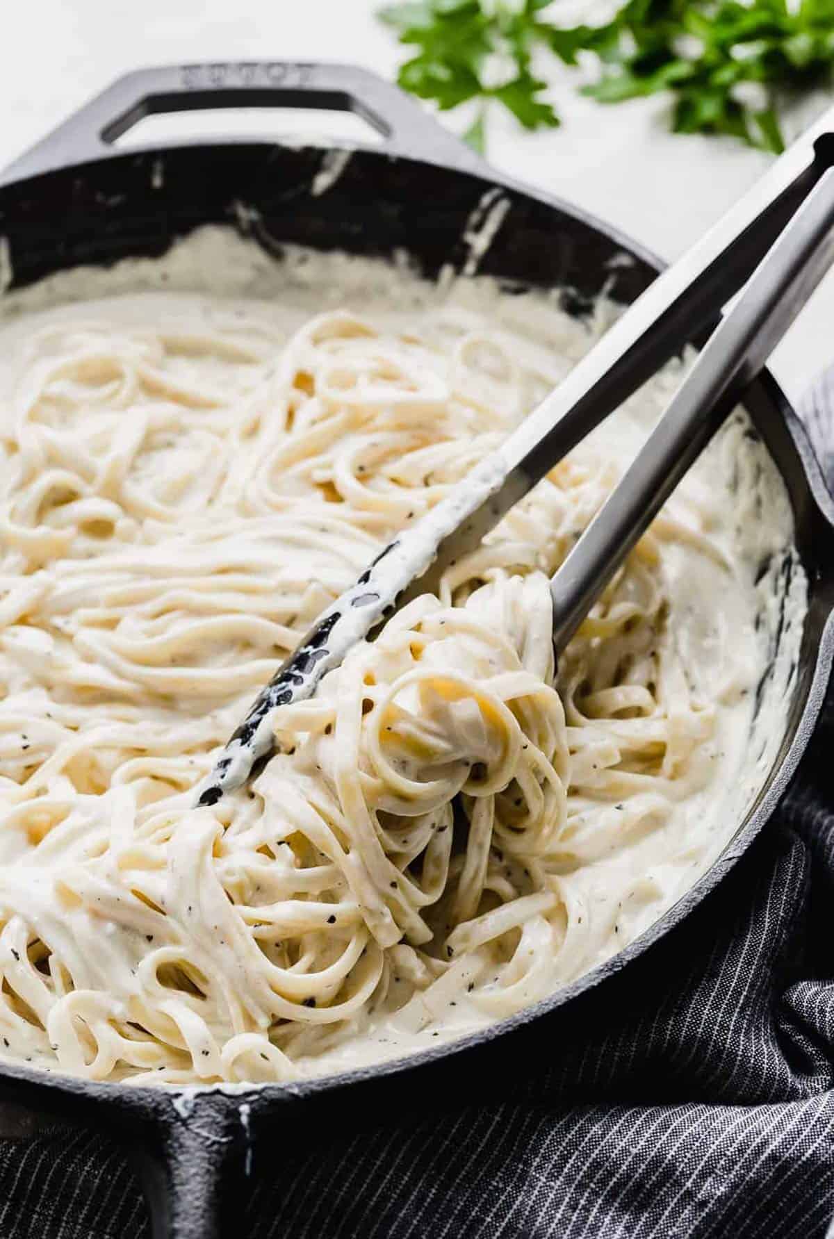 Kitchen tongs stirring creamy Fettuccine Alfredo in a black skillet.