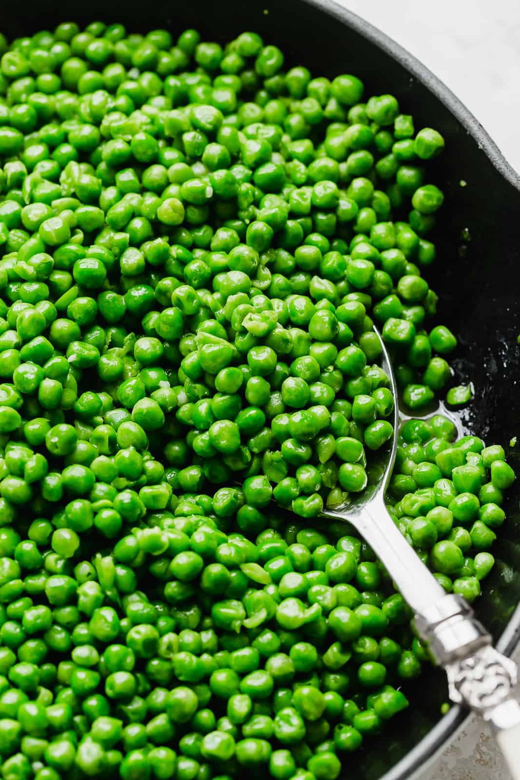 A serving spoon scooping up freshly cooked green peas from a skillet.
