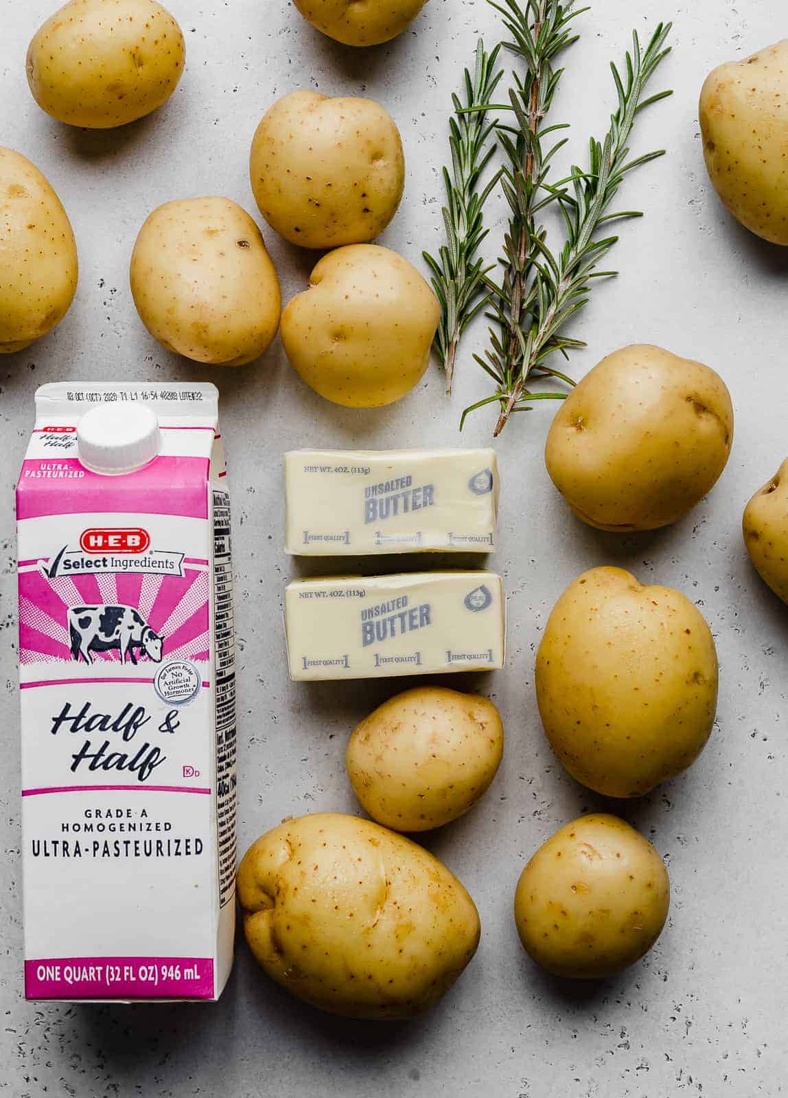 Ingredients used to make mashed potatoes: potatoes, half and half, butter, and rosemary against a grey background.