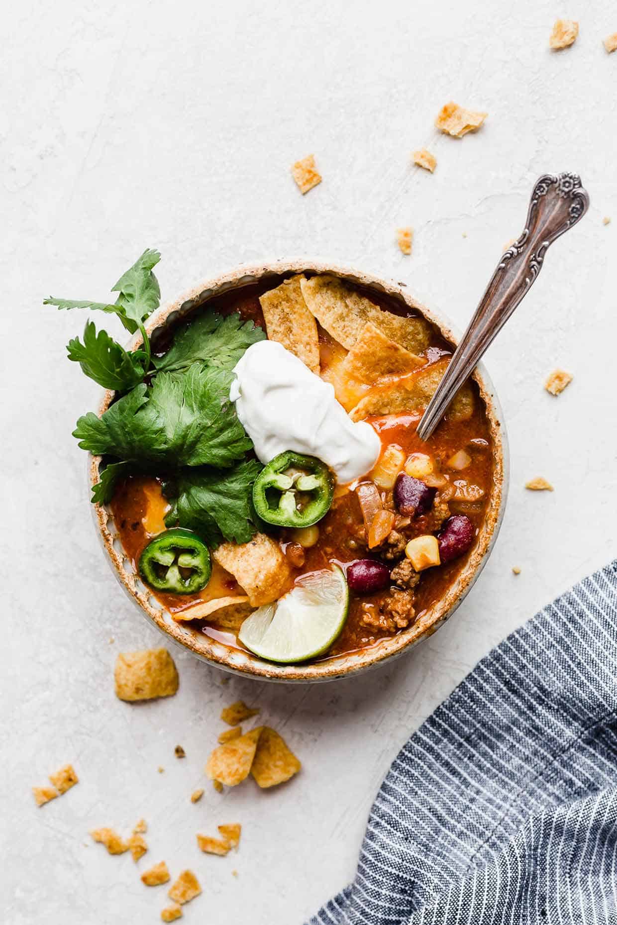 A bowl on a white background, filled with Taco Soup with Fritos, sour cream, cilantro, and sliced jalapeños.