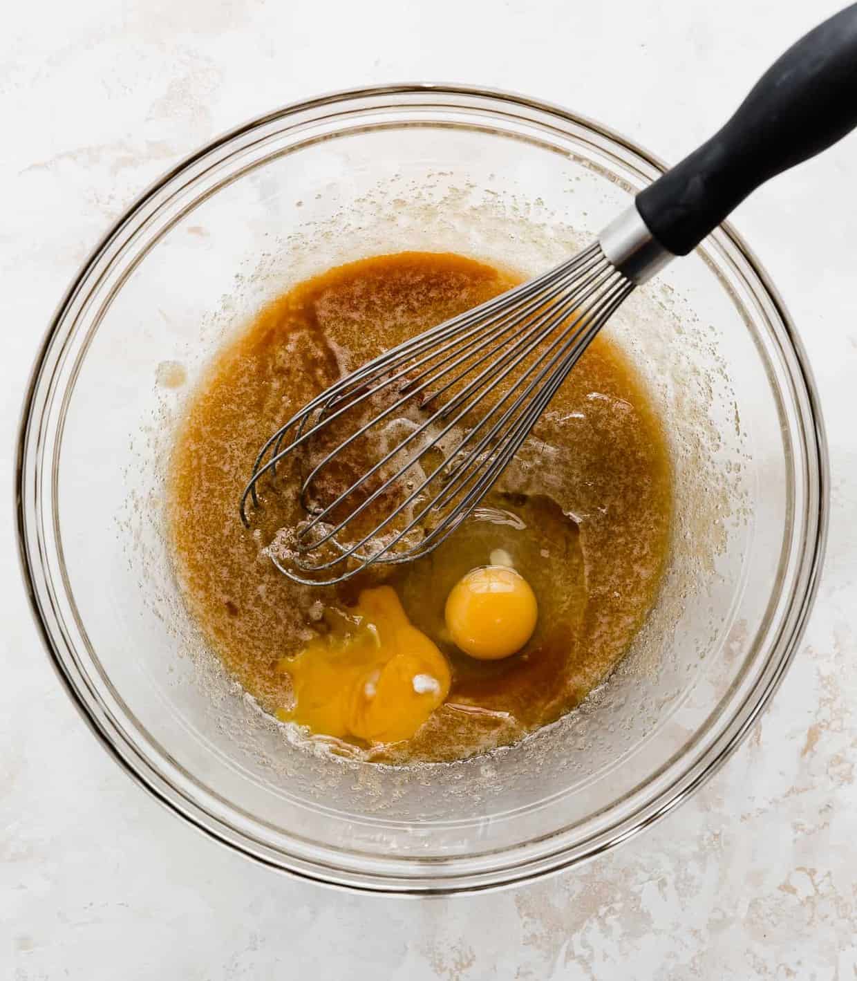 Brown sugar, melted butter, eggs, and vanilla in a glass bowl being mixed by a whisk.