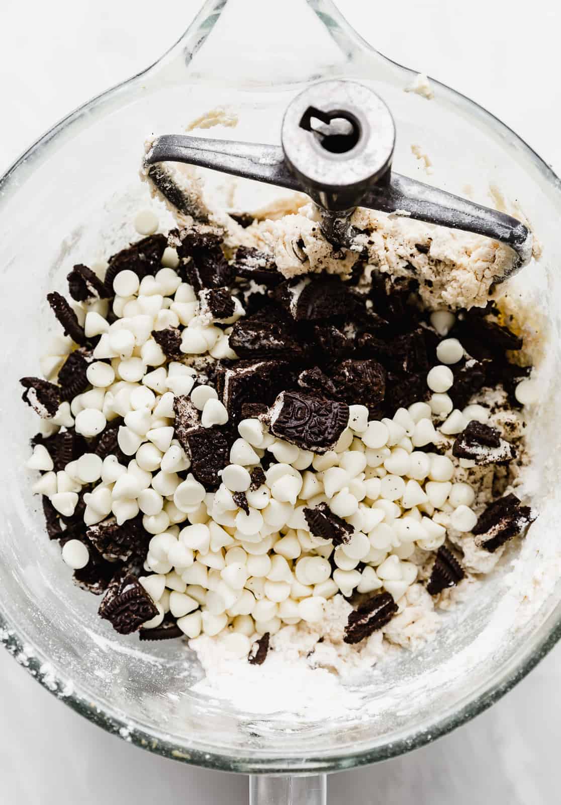 A large glass bowl full of Cookies and Cream Cookie dough batter with white chocolate chips and chopped Oreo's on top.