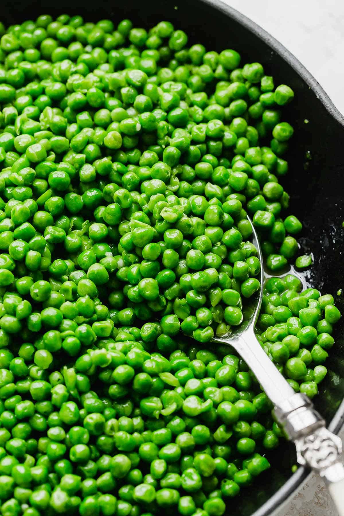 A silver serving spoon scooping up peas from a skillet.