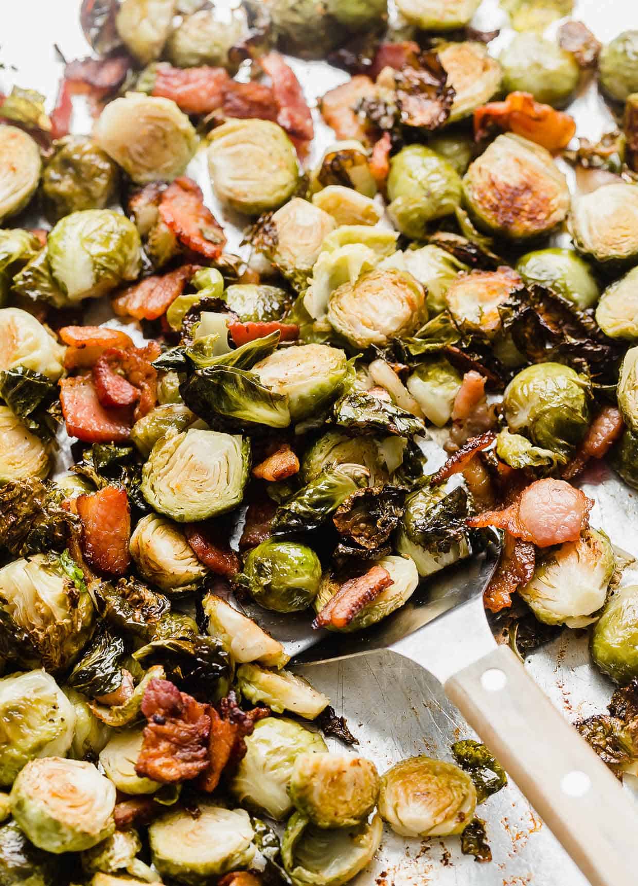 A metal spatula scooping off Roasted Brussels Sprouts with Bacon from a baking sheet.