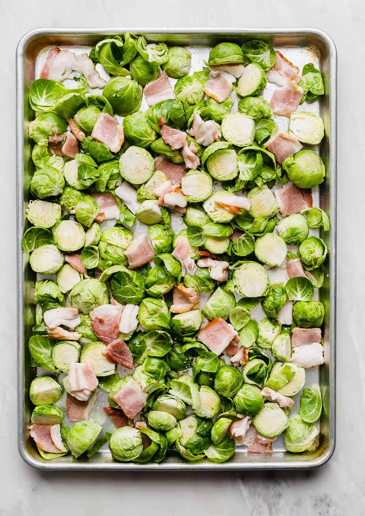 Unbaked Brussels sprouts and raw bacon chunks on a baking sheet.