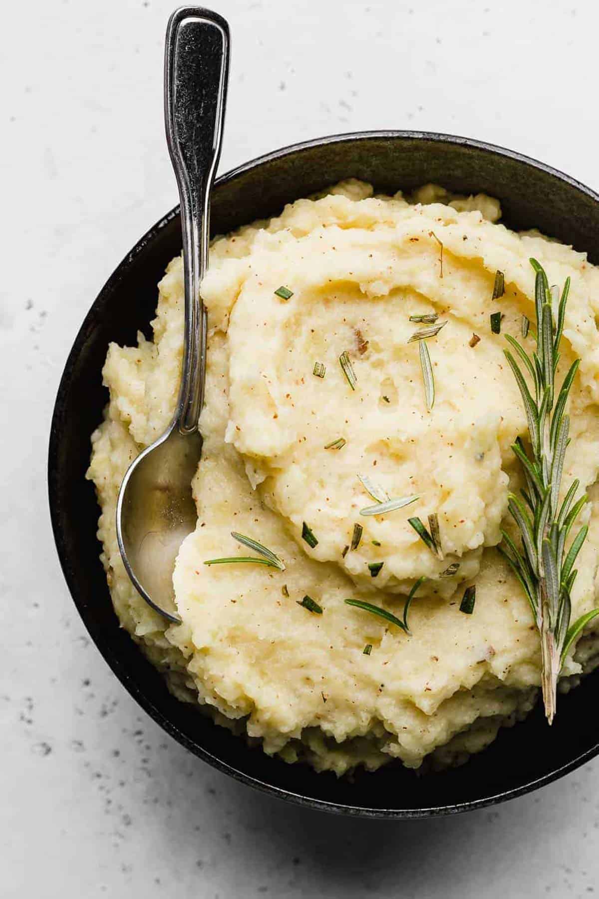 A spoon nestled in a black bowl that's filled with creamy Rosemary Brown Butter Mashed Potatoes.