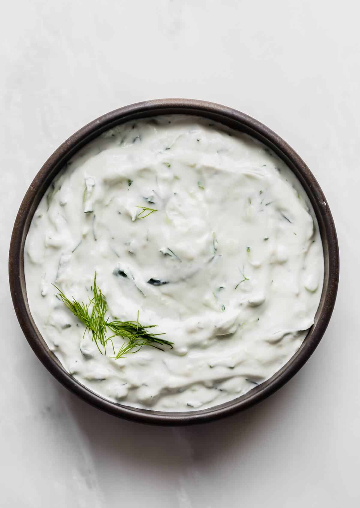 A black rimmed bowl filled with authentic Tzatziki sauce using shredded cucumber and fresh dill, on a white background.