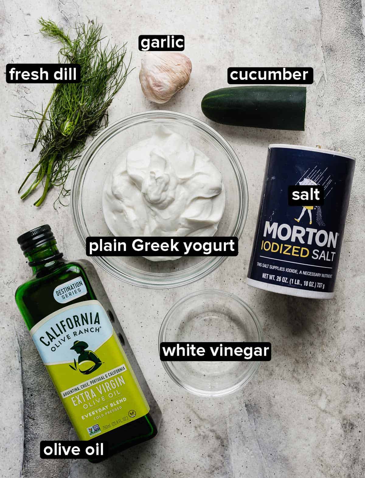 Tzatziki ingredients on a light gray background: salt, cucumber, dill, olive oil, greek yogurt, garlic, and vinegar.