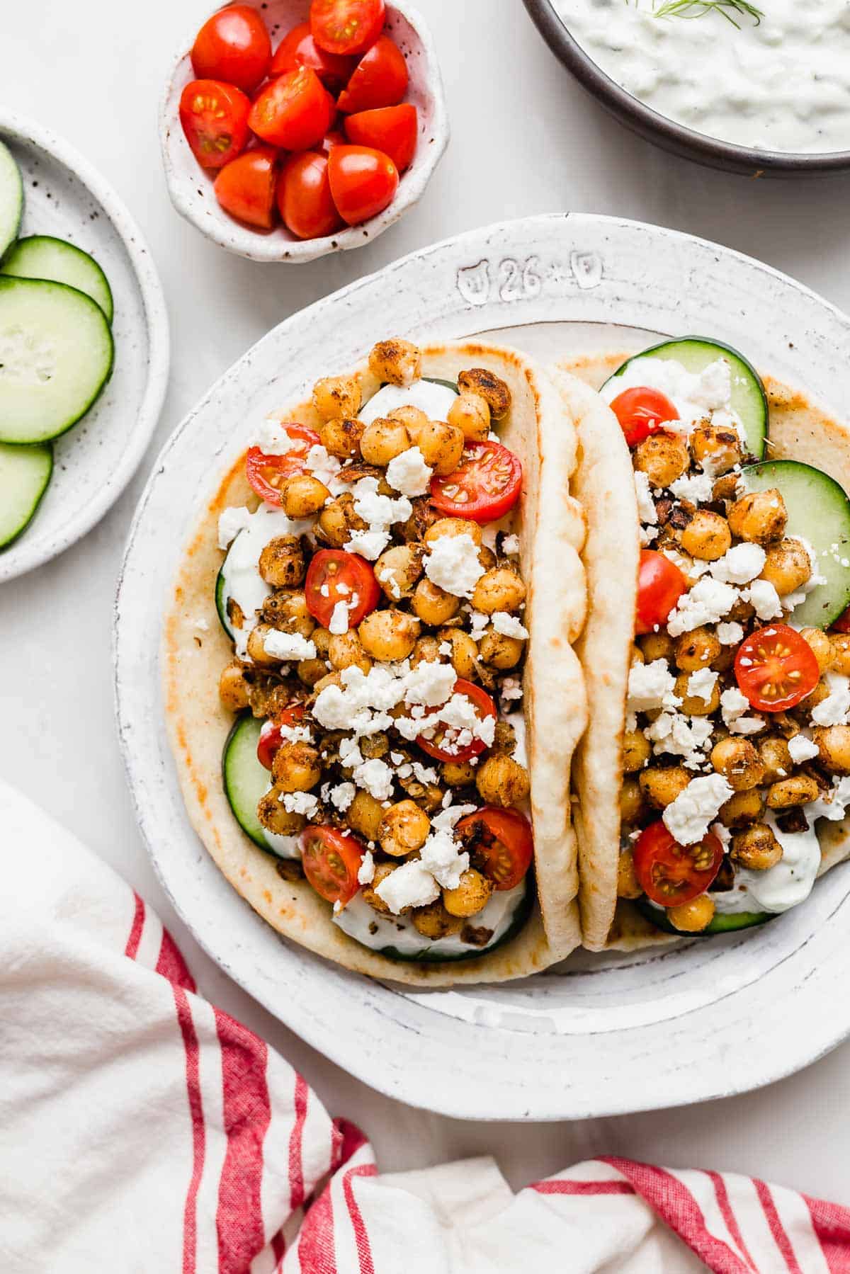 Two Chickpea Gyros on a white plate with a small bowl of cherry tomatoes, sliced cucumbers, and tzatziki sauce towards the top of the image.