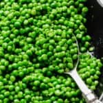 A silver serving spoon scooping up properly cooked frozen peas from a skillet.