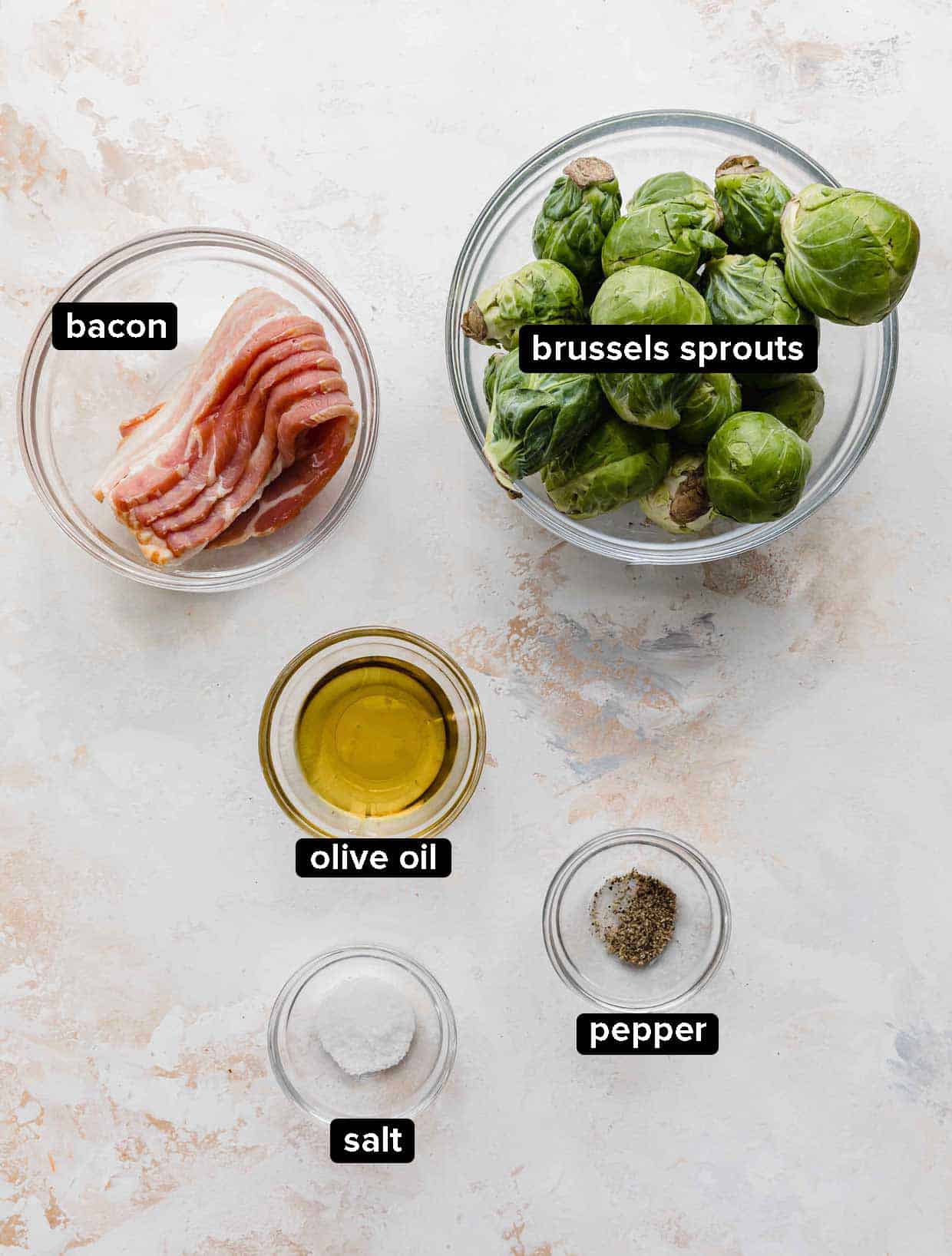 Roasted Brussels Sprouts with Bacon ingredients portioned into glass bowls on a white background; bacon, Brussels sprouts, olive oil, salt and pepper.